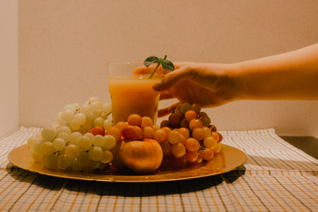 Faceless woman's hand reaches for a glass of freshly squeezed fruit juice on a tray among various seasonal tropical summer fruits. Still life nostalgic retro effect warm colors. Refreshment drink foodの写真素材