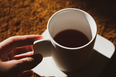 Stylish white ceramic or porcelain tea, coffee cup in woman's hand. Girl drinking a hot beverage from a beautiful crockery holding a mug in hand. Female hands in the shadow. Cozy morning scene.の写真素材