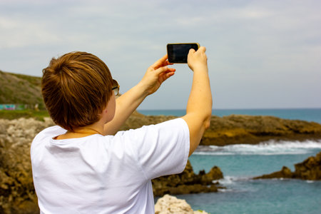 Middle age woman takes a photo on mobile phone on the Atlantic coast traveling through Spain, Santander. Female tourist with a short haircut in casual clothes on a seaside, seashore. Summer vacations.の写真素材