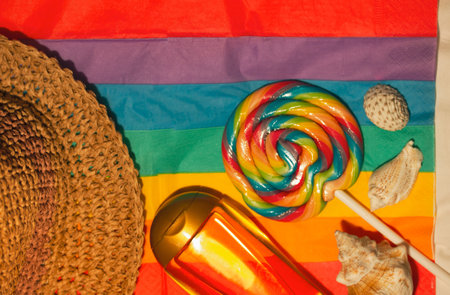 Straw hat, multicolored lollipop, suntan oil bottle on bright rainbow background. Accessories for summer vacations on sea, ocean shore. LGBT, gay symbol, pride month. Summertime fun joy photography.の写真素材