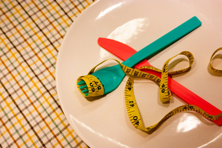 White plate on checkered tablecloth. Measured yellow tape, colorful plastic utensils. Serving the table during the diet, therapeutic interval fasting. The concept of weight loss, tracking weight loss.の写真素材