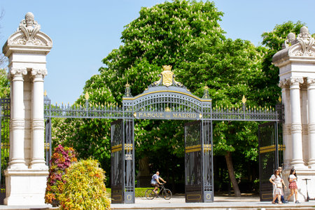 Madrid, Spain. 7 May 2022. Black metal gate with white columns at entrance to Retiro Park. Botanical Garden on a sunny spring day when the trees are in bloom. People walking outdoors, riding bicycles.のeditorial素材