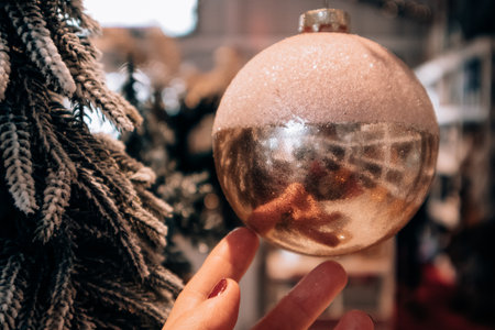 Large glass golden pink ball in woman's hand on blurred background. Christmas tree toy background. Interior decor for New Year's Eve 2023. Choosing toys for evergreen New Year pine tree in the shop.の写真素材