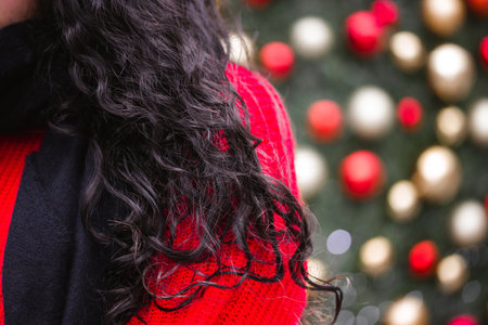 Young woman, brunette girl with dark curly hair, beautiful curls in red sweater on blurry background of Christmas tree with bright Christmas tree toys. New Year 2023, winter holidaysの写真素材