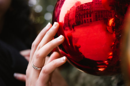 A young woman, girl reaches out and touches with a hand a red glass ball, Christmas tree toy. New Year's Eve 2023. Christmas magic, wintertime festive background. A silver ring onの写真素材
