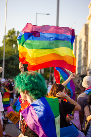 Madrid, Spain. 26 July 2022 Lesbian girl holding a developing rainbow flag, symbol of LGBT community at a gay parade in the summer. Pride month party celebration on the city street. People with flag.のeditorial素材