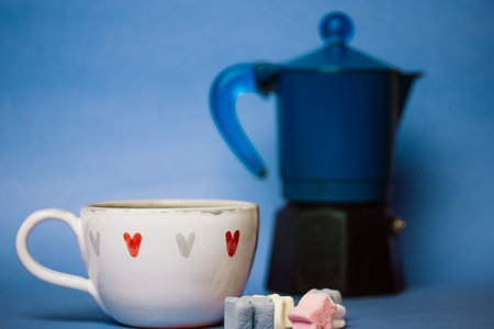Small white cup in hearts with espresso coffee, cocoa or hot chocolate drink on blue background. Marshmallows on a table. sweet dessert. Cozy still life with drinks. Geyser coffee maker on foreground.の写真素材
