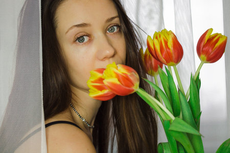 Beautiful young white woman with blue eyes portrait close-up with a bouquet of orange yellow tulips. Girl sniffing blooming bulb lowers indoors. congratulations for women's day. Mysterious calm ladyの写真素材
