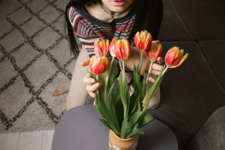 Young woman indoors puts a bouquet of spring bulb flowers in a vase in a domestic apartment, cozy home. girl sniffing orange tulips. A armful of flowers in a woman's hands. Beautiful homy atmosphereの写真素材