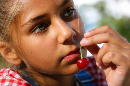 A little teenage girl holding a ripe red cherry in the fruit trees orchard. Portrait of a beautiful oriental tanned child, female kid 10-12 years old in summer nature. Natural hippie children beauty.の写真素材