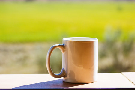 White cup mug of tea or coffee standing on windowsill against green grass outside window on sunny warm day. A morning drink without people on a nature in summer. selective focus. Relax, lounge conceptの写真素材