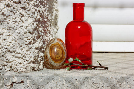 Easter egg with natural stone texture. Spring still life with a blooming tree branch in a small red glass vase against white wall background. Cinematic vintage authentic Easter holiday photo.の写真素材