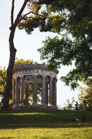 Madrid, Spain. September 1, 2022 Picturesque Capricho Park with an ancient antique stone gazebo on top. Spanish sculptures and landmarks. Botanical garden pavilion. People resting on a grass in summerのeditorial素材