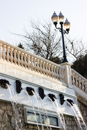 Fencing on an antique white stone marble fence in city park with a cascade of artificial streams of water, waterfalls, fountains. Metal black street lamp Vertical background with architectural detailsの写真素材