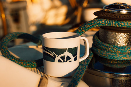 White cup with drawn helm on deck of a white yacht, ship, boat. Concept of tea coffee drinking while traveling at sea ocean. A mug among the sea ropes in morning sunshine light. Nautical wallpaper.の写真素材