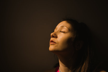 A face of a young beautiful woman girl with eyes closed, exposing face to the warm sun light. Dreaminess, fantasy, anticipation concept. Portrait in profile with space for text on a dark background.の写真素材