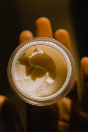 Round jar of face skin care cream in woman's female hand on natural brown background top view. Beauty treatments, self-care concept. Beautician procedure professional. Lotion container Dark key photoの写真素材