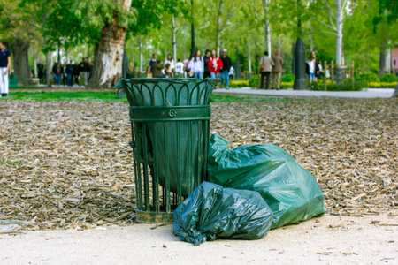 Green metallic trash can on the street in a city park outdoors. Organic eco garbage bags filled with all kinds of garbage on a pavement. Rubbish bin on nature. Clean environment, ecology concept.の写真素材