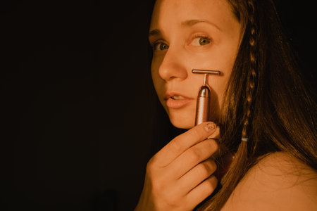 Young beautiful white woman, girl is using vibrating golden facial massager on dark black background...