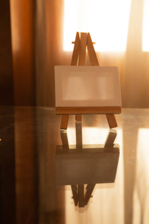 White canvas on wooden easel on a glass table vertical photo. School of drawing, creativity, creating pictures, works of art, masterpiece concept. creative crisis. Warm lighting indoors in a room.の写真素材