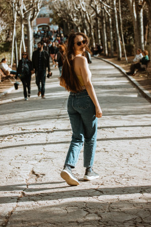 A beautiful European white brunette girl with long brown hair in jeans is walking outside in a city park in a summer sunny day. People in a street. Back view of a young woman going awayの写真素材