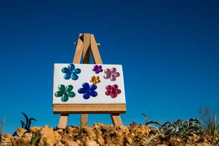 Wooden miniature easel with shiny artificial multicolored blooming flowers. Creating a painting with a floral composition in nature. Hobby, leisure. Drawing school lesson. Art and creativity concept.の写真素材
