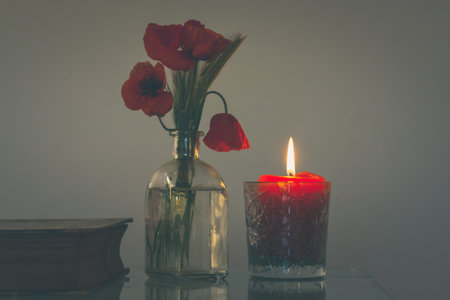 Dark still life with a bouquet of red poppies in a vase, an old book, a burning candle on the table. Decor for a cozy holiday home. Exploring spirituality, wisdom, knowledge concept. wild flowers.の写真素材