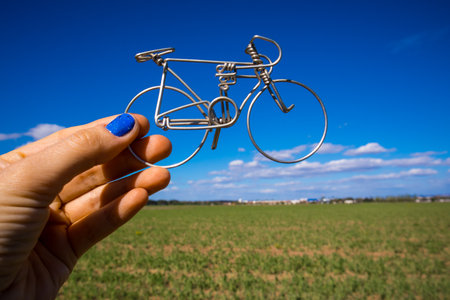 A metal bicycle in a woman's hand against a blue sky in summer day Holding a toy mini bike. Cycling outdoors. Round aluminum wheels of a bike. Healthy lifestyle, leisure, sports activity. holiday tripの写真素材