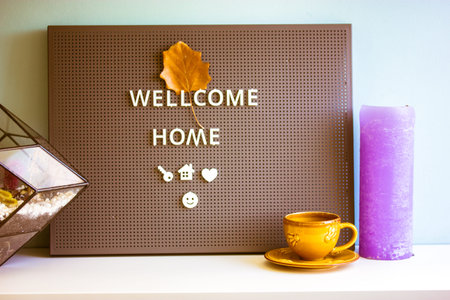 Black decorative message board with text Welcome home, autumn orange leave. Modern interior of a residential apartment. Yellow ceramic coffee cup and purple candle. Sweet cozy home, hygge.