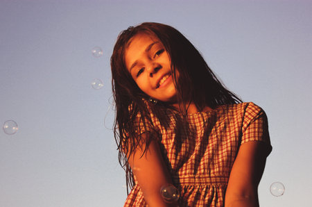 Cute beautiful happy teenage girl 10-12 years old in the background of the sky. Beautiful tanned teen child, kid smiling standing among soap bubbles in the air. happy childhood. Summer holidays fun.の写真素材
