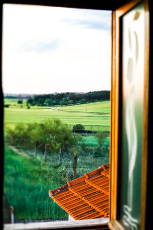 Vertical photo of an open window in a rustic house overlooking green meadows with fresh grass on a spring summer day. Authentic landscape. View from a window on a street, nature. Rural scene.の写真素材