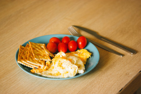 A blue plate of fried eggs, cherry tomatoes, croutons on a wooden table in a rustic kitchen. Healthy food eating, protein and vegetables. Dish served on the table, setting for a meal. A breakfast, lunchの写真素材
