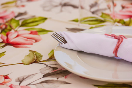 Elegantly table set at wedding, christening banquet. Floral tablecloth with pink flowers and white round plate, napkins. Thanksgiving holiday decor. DIY napkin for birthday party lunch, brunch.の写真素材