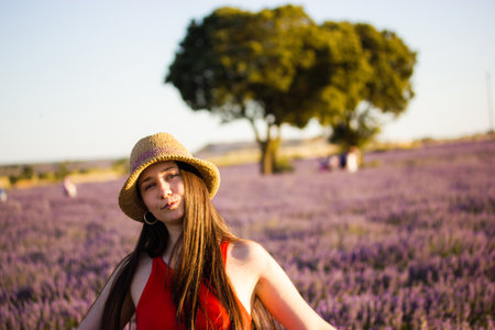 A beautiful romantic brunette young woman wearing a straw hat in a blooming lilac lavender field. Female portrait in nature on a summer day. Traveling in Europe. Boho style clothing. Provence, France.の写真素材