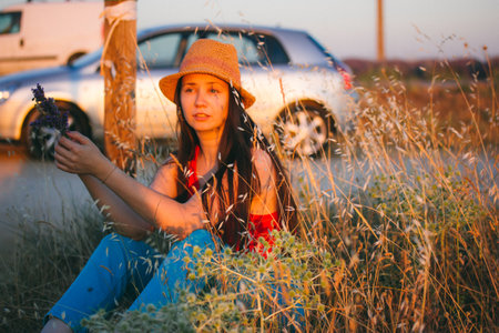 A young brunette girl wearing a straw hat sits in a meadow, a field on a summer day. Traveling by car through the countries. Interesting life, adventure in the nature. A woman among wildflowers.の写真素材
