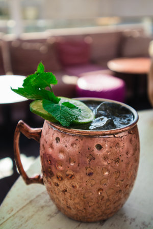 Alcoholic cocktail with a slice of lime, mint leaves, cinnamon, ginger. Serving of drink on a wooden table in a cafe, bar. Cold frosty moscow mule cocktail in bronze golden copper mug close up detailsの写真素材