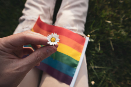 White camomile, daisy flower in female hand and colorful LGBT flag in rainbow colors on background. A lesbian, bisexual, gay girl sits relaxing in green clearing in park on sunny day. Pride Month.の写真素材