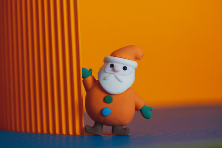 Toy miniature plasticine jolly Santa Claus waving on a bright orange and purple background. Winter Vacation. Holiday wallpaper New Year 2024. Merry Christmas postcard with copy space. Little figurineの写真素材