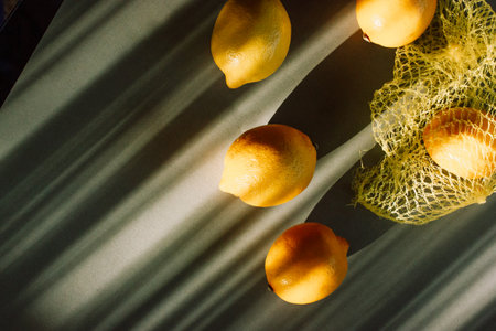 Yellow ripe lemons fall out of the ecological bag left long diagonal shadows on a green table. Trendy stylish modern food background. Yellow net shopping bag with citruses in sunny summer day.の写真素材
