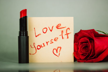 Red lipstick next to a note written in lipstick with a text 'Love yourself' and a heart shaped draw on a woman's cosmetic table. Motivation, narcissism, romance concept Red rose from admirer on a dateの写真素材