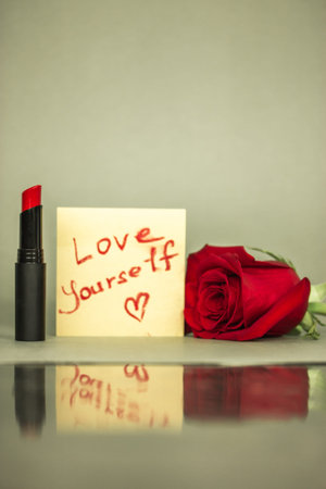 Red lipstick next to a note with hand written text 'Love yourself' and a heart shaped draw on a...