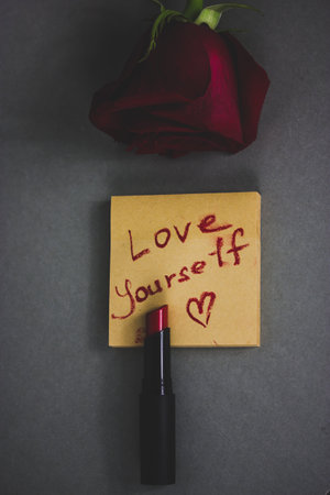 Red lipstick and a note with hand written text 'Love yourself', a heart shaped draw on woman's dark cosmetic table. Motivation, narcissism, romance. A single rose for lover on Saint Valentines Day.の写真素材