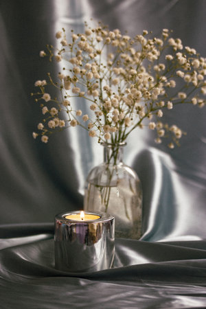 A burning candle in a metal candle holder on a table covered with a silver silk. Luxurious stylish decor for modern interior. A glass vase with small white summer flowers indoors. Floral postcard.の写真素材