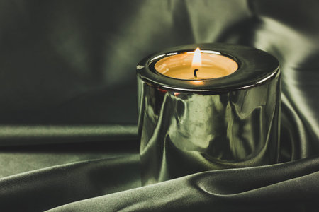 A candle in a metal holder on a dark olive green silk tablecloth. Stylish decor.
