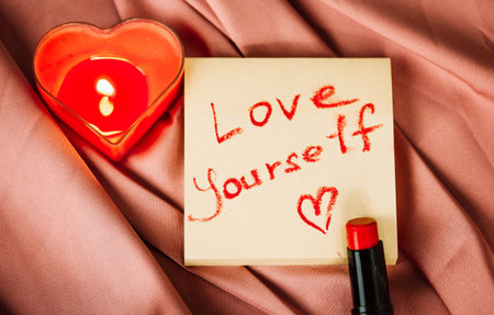 A note to self with a call to love yourself drawing with red lipstick on a silk terracotta cloth top view. A burning heart-shaped red candle, interior decor for womens home. Happy Valentines Day 2024の写真素材