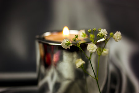 Spa still life with white small fresh blooming flowers, metallic burning candle on silk silver gray background. Wedding accessories, floral arrangement, blooming sprig from bouquet. Relaxing at home.の写真素材