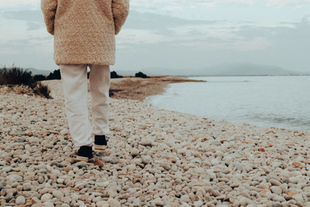 Young woman in a stylish white coat luxury style strolls along the sandy coast in fall cold day. Autumn landscape, seascape. Female traveler solo.の写真素材