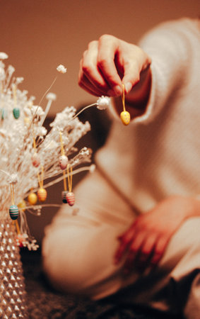 A woman hangs a yellow decorative Easter egg on a bouquet of dry white flowers in the interior. Festive home decor. Woman's hand holding toy pendant.の写真素材