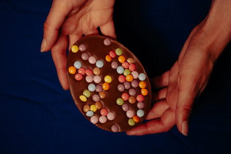 Large chocolate Easter egg with colorful candies in female hands Dark table top view. Easter's...