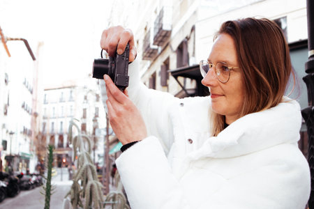 Madrid, Spain. 1 March 2024. A young woman in a white fur coat takes a photo with a small digital camera on a city street. Female travel photographer.のeditorial素材